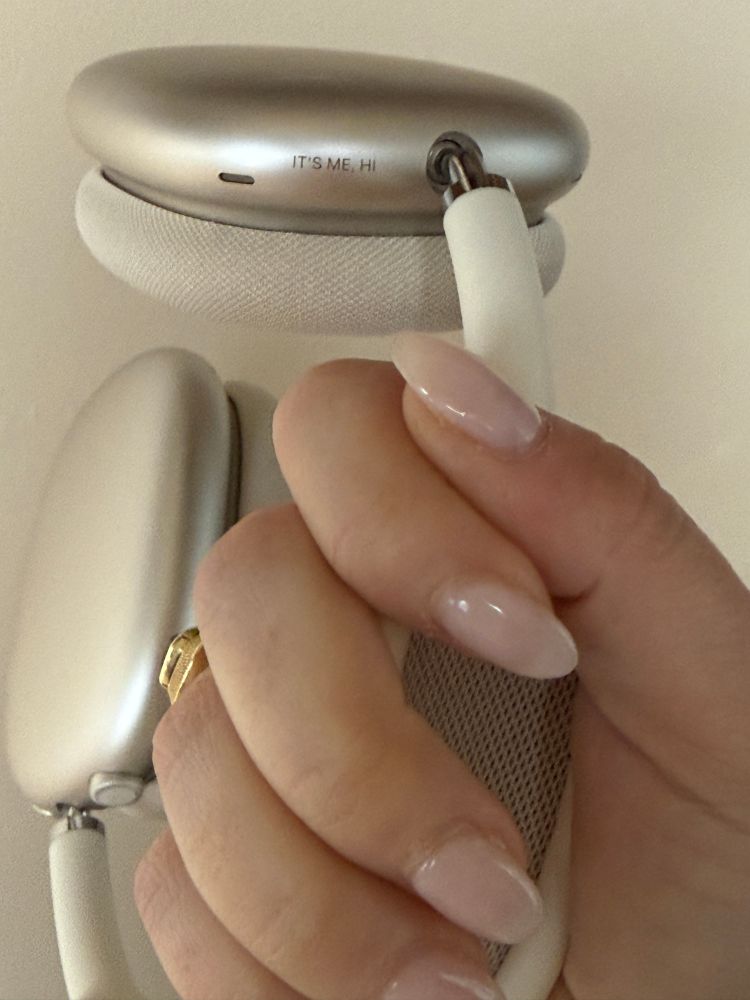 a white woman’s hand with light plink nails holding white airpod max headphones engraved with “IT’S ME, HI”