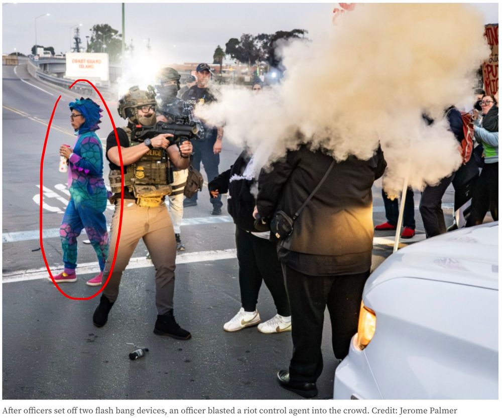 The above picture of an ICE agent in tactical gear who is firing flash bangs at the heads of two non violent protestors only a few feet away. the ICE agent has clearly peed his pants. Behind him, a person in a fleece blue onesie with a hat that looks like a dinosaur is walking with their water bottle.