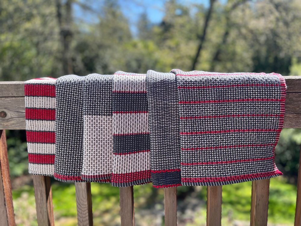 6 tea towels are folded in half and draped over a wooden railing. Each towel is a different pattern but all are some combination of white, dark gray, and red.