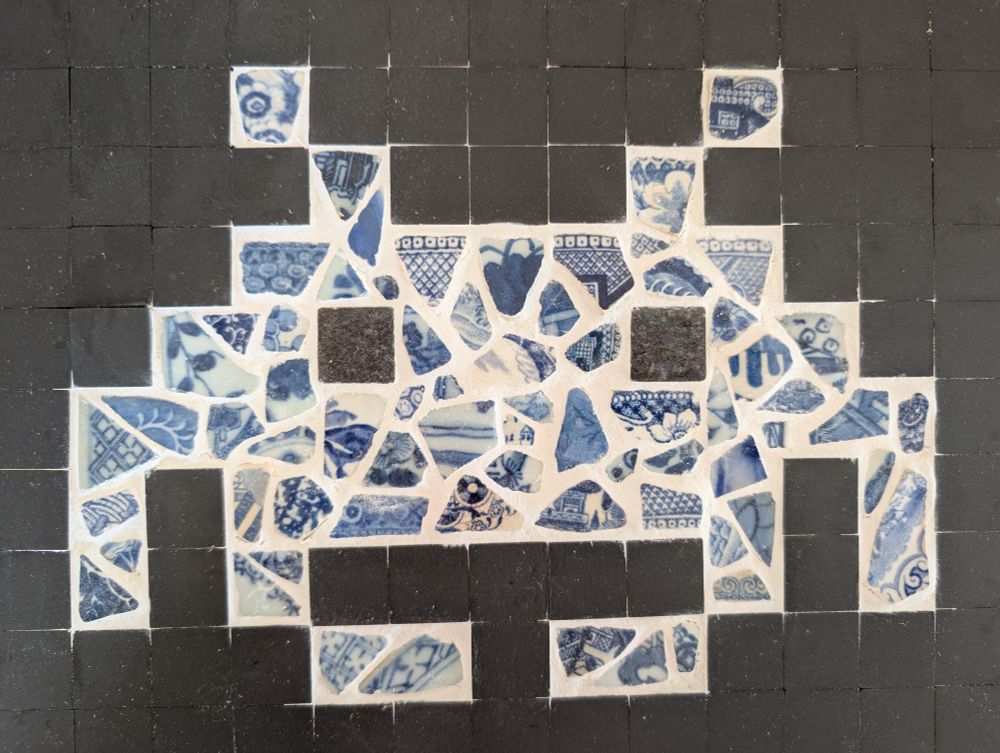 A mosaic in the shape of a space invader with black tiles on the outside and blue and white pottery sherds and white grout.