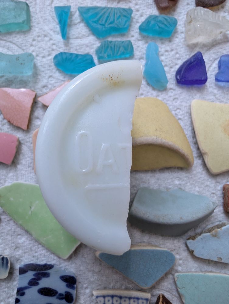 A piece of white broken pot that says "Oat" on it, with a background of other colourful pieces of glass and pottery sherds.