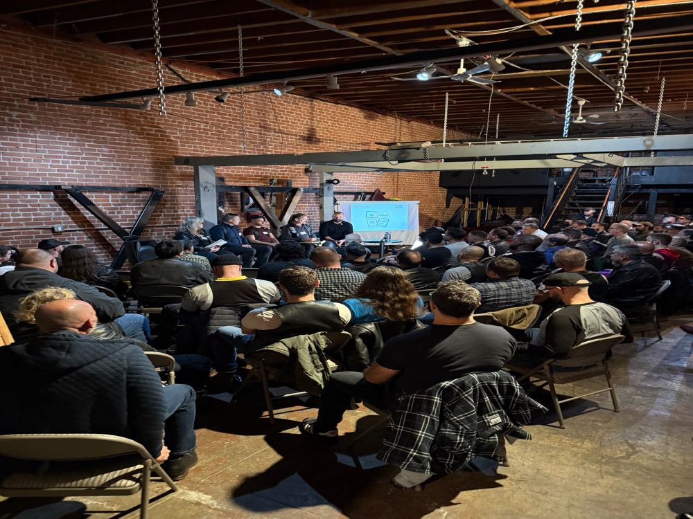 75 people sitting in chairs in a community dungeon space listening to 4 presenters seated at the front of the room