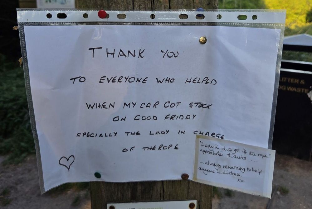 Shows a hand written notice that is pinned to a post. Notice is written in marker pen and reads "Thankyou to everyone who helped when my car got stuck on Good Friday. Especially the lady in charge of the rope."
A smaller sign below is written in different handwriting and reads "Lady in charge of the rope appreciates the thanks. Always rewarding to help anyone in distress."