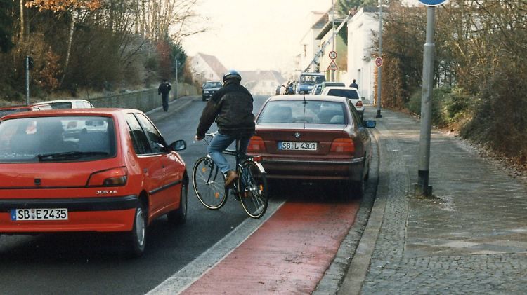 Foto: Auto auf einem Radweg zwingt Radfahrer zum Ausweichen auf die Straße , wobei er vor den Kühler eines Autos kommt.