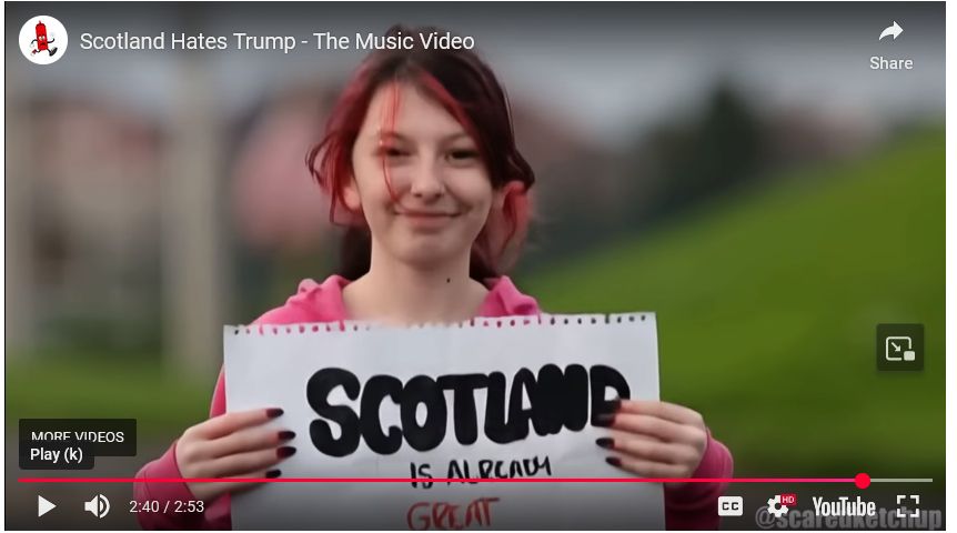 Still image from the end of the music video “Scotland Hates Trump”: a confidently smiling girl with partially dyed black and red hair and black fingernails holds a sign with a handwritten messag: "SCOTLAND IS ALREADY GREAT"