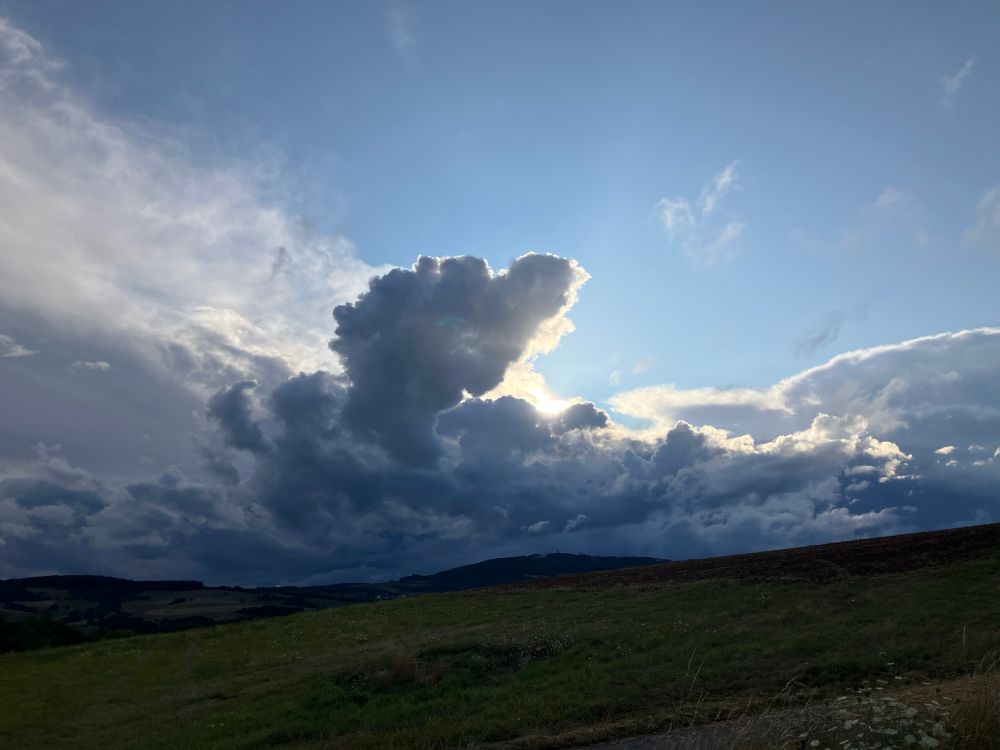 Clouds floating over fallow fields in beautifully green Rheinland Pfalz, with the sun shining behind them. The sky is pale blue behind them, and they have many shades of grey