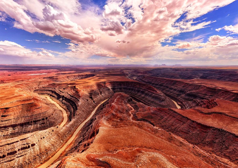 This photo depicts a stunning desert landscape featuring deep, winding canyons carved by a meandering river. The canyon walls are layered with shades of red, orange, and brown, showcasing the rich geological history of the region. The river below reflects a muted, golden hue, contrasting with the vibrant earth tones. Above, a vast expanse of sky is filled with dramatic, fluffy clouds that scatter sunlight across the terrain, enhancing the textures and colors. The horizon stretches endlessly, blending the rugged land with distant mesas and plateaus, creating a breathtaking and serene vista.
