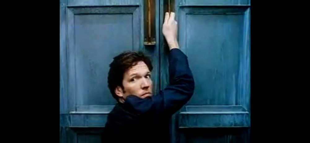 Martin Donovan in Amateur. He is looking back over his shoulder as he moves to open one of two enormous blue double doors that are approximately Tardis in color.