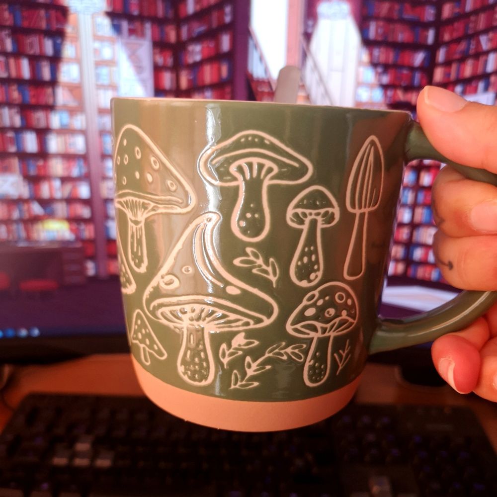 Una mano sujeta una taza verde musgo con setas grabadas en líneas blancas con un fondo de pantalla detrás que es una biblioteca.