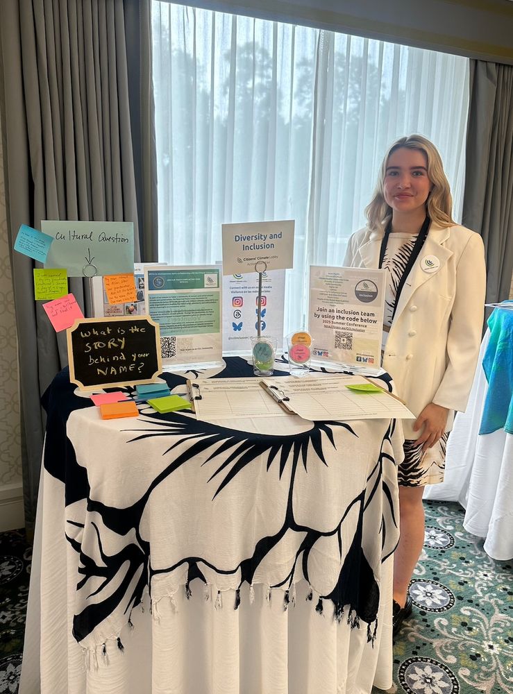 A white woman in her 20s stands behind an informational booth, which contains information about diversity and inclusion at Citizens Climate Lobby.