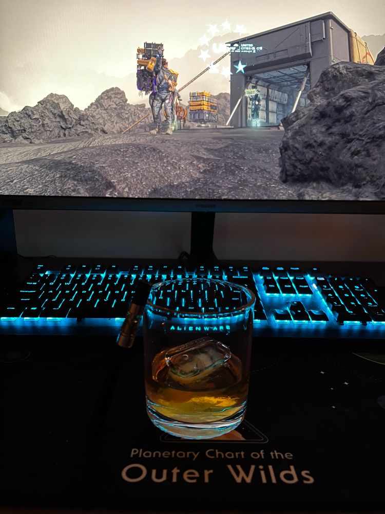 A glass of whiskey with a vape laid against it. Sitting in front of a keyboard and monitor with an image of Death Stranding.