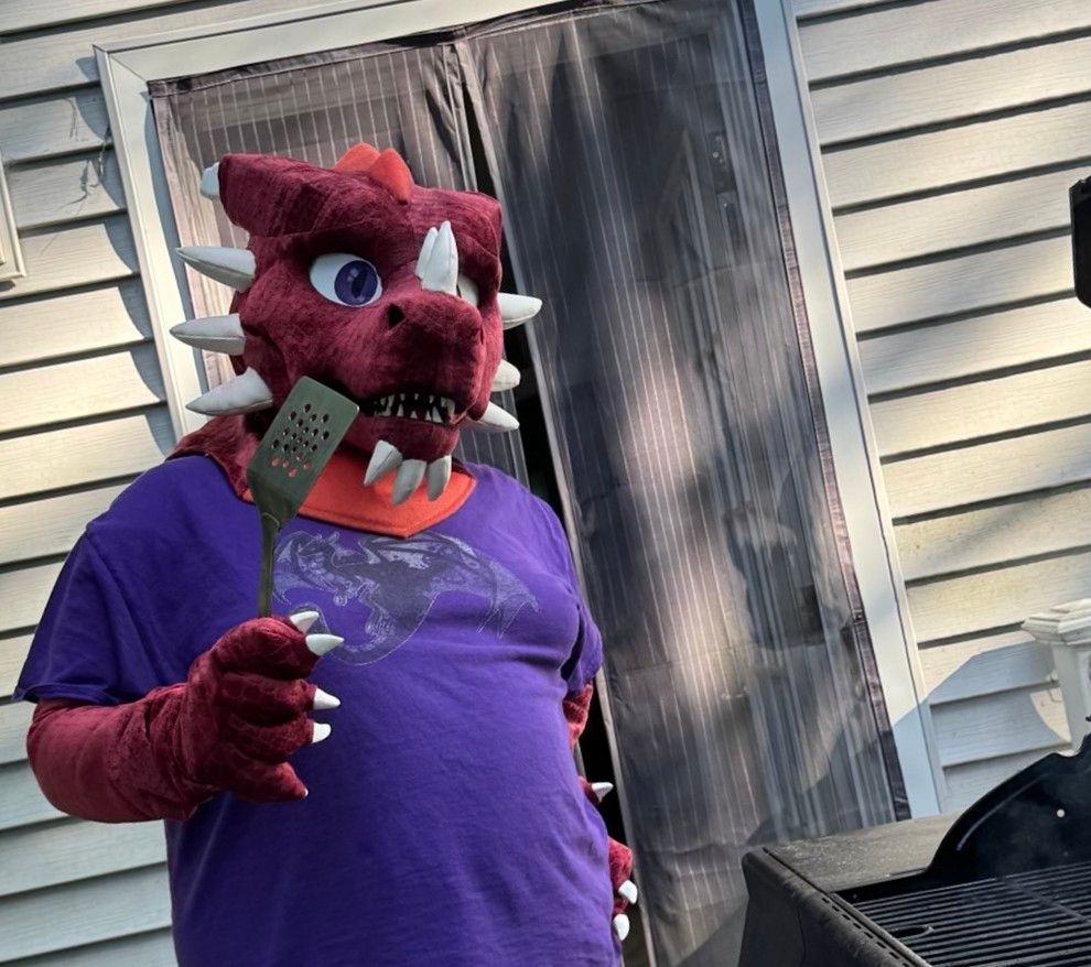 Red dragon fursuiter holding a spatula in front of an outdoor grill