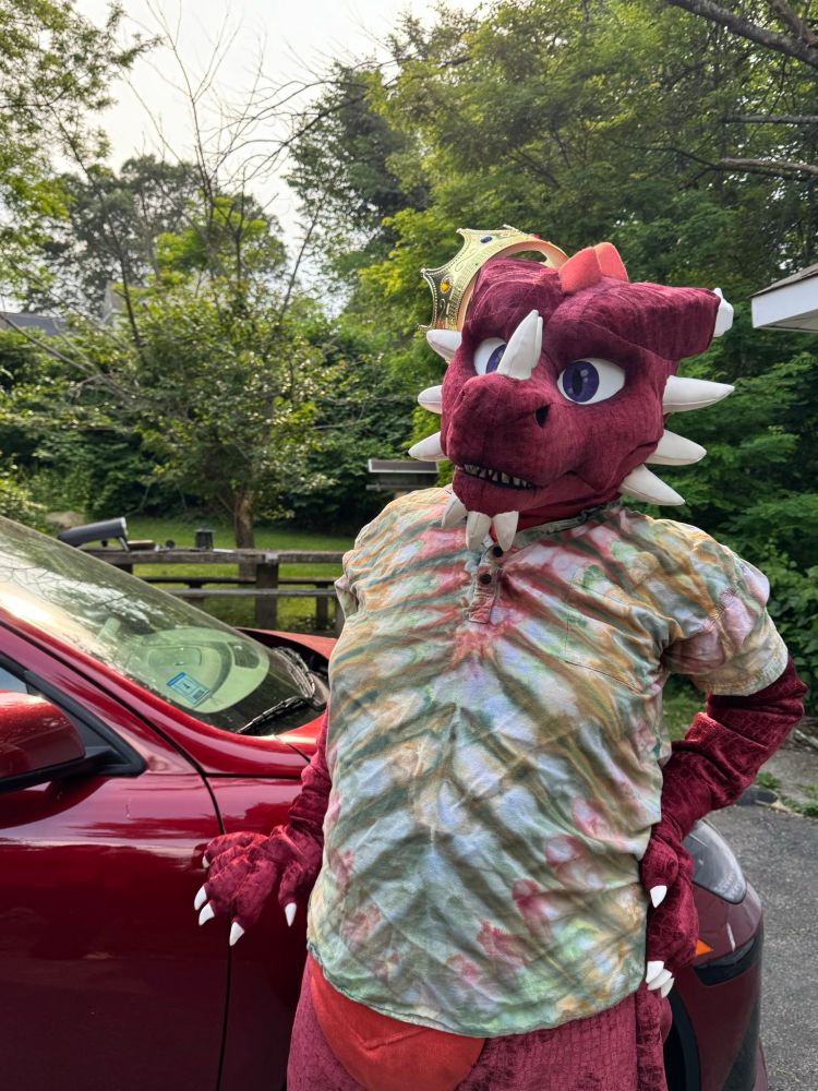 A red dragon fursuiter wearing a shirt leaning on a car