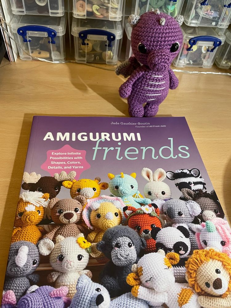 Photo of a book - Amigurumi Friends by Jade Gauthier-Boutin. The title and author are at the top and the purple cover is full of cute crocheted animal characters. The tag says ‘20 easy patterns to create 100+ adorable critters’. Next to the book, at the top, is a small purple crocheted dragon with grey wings and spikes.