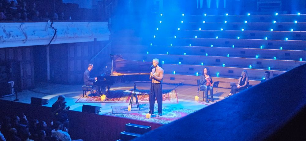Shows musician Teeks on stage,  next to a grand piano. Violinists behind him to the right