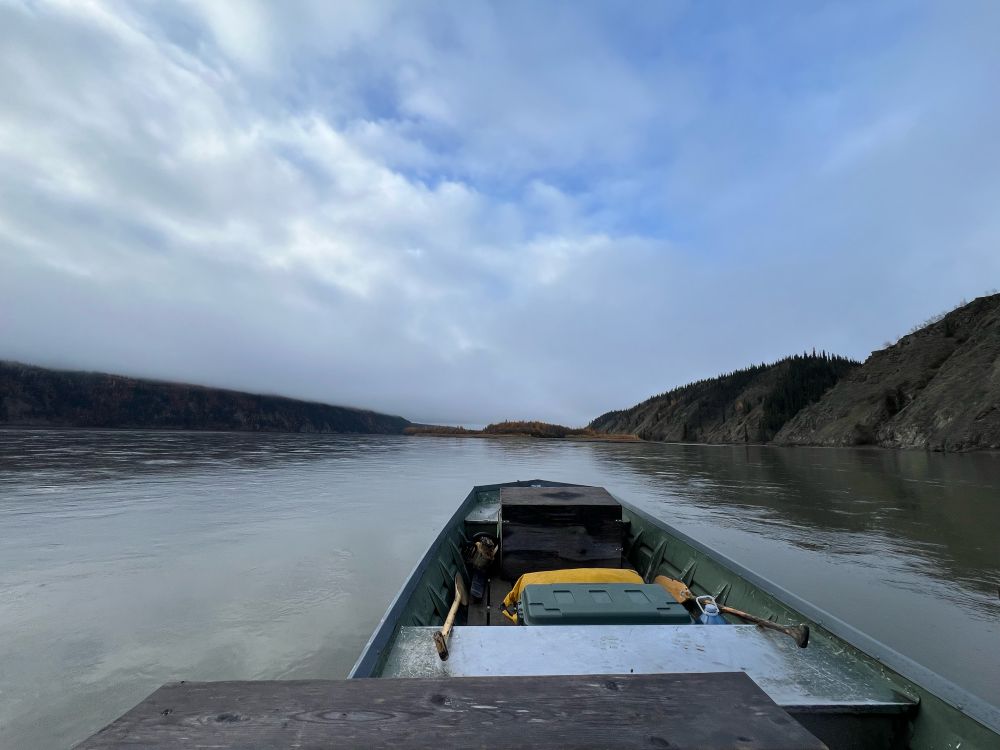 Wide angle view from inside a river boat. Water and rocky treed hills on all sides, paddles, bins, etc inside the boat. Sky has low clouds with blue above. 