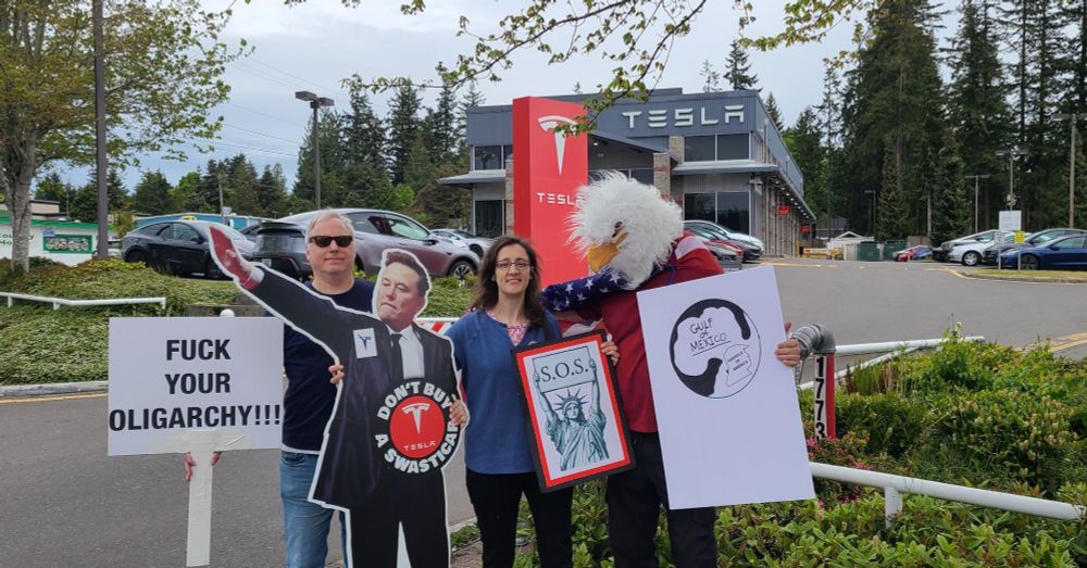 A small cluster of protesters in front of the showroom signage; the most visible sign says "Fuck Your Oligarchy!", and another has a picture of the Statue of Liberty holding up an SOS sign.