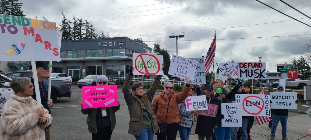 Photo of folks lined up in front of the Tesla showroom in Lynnwood with signs like (F)elon, Send Elon to Mars, and Defund Musk.