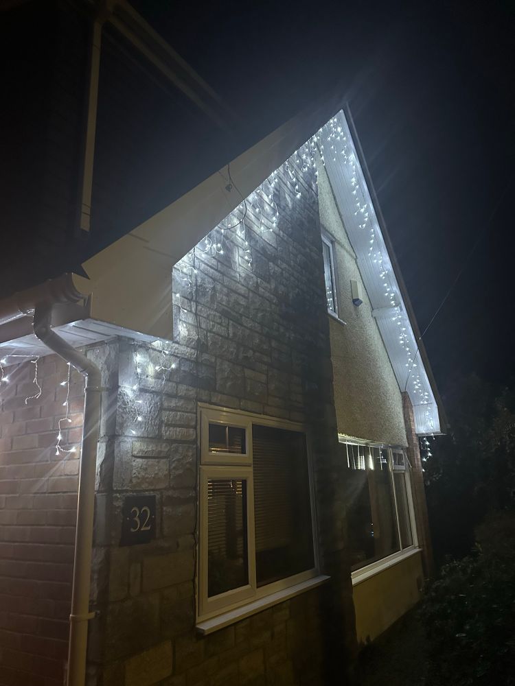 A photo of the front of a dormer style house. It’s taken in the dark to show the Christmas lights, which have been hung along the edge of the roof. The lights have short dangling icicles along them. 