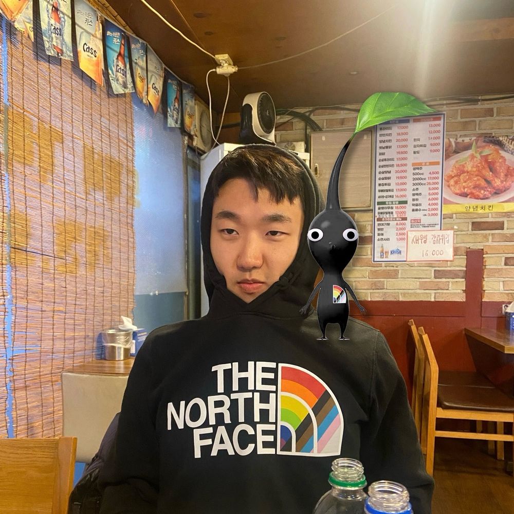 노란색 노스페이스 프라이드 후드를 입은 별이와 그를 닮은 검은색 피크민 캐릭터가 그의 어깨 위에 있는 사진