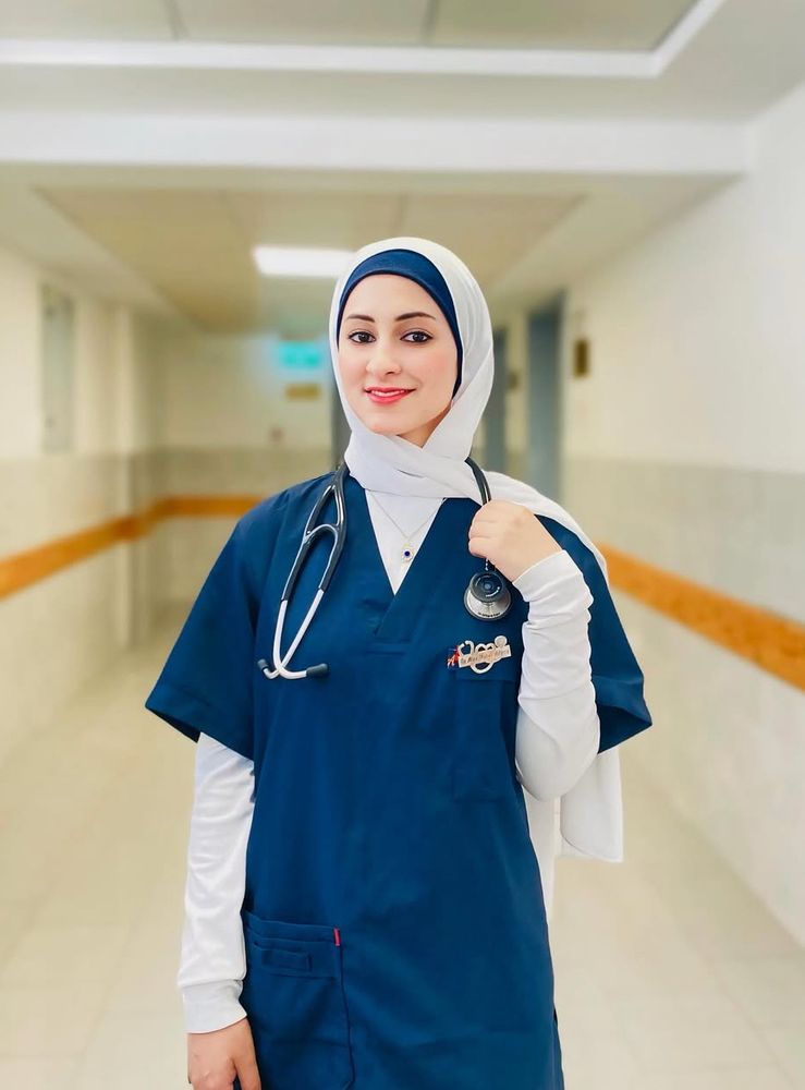Alaa stands smiling in the corridor of a hospital, wearing navy blue scrubs with a white long sleeve underneath, white hijab and and a stethoscope across her shoulders. 