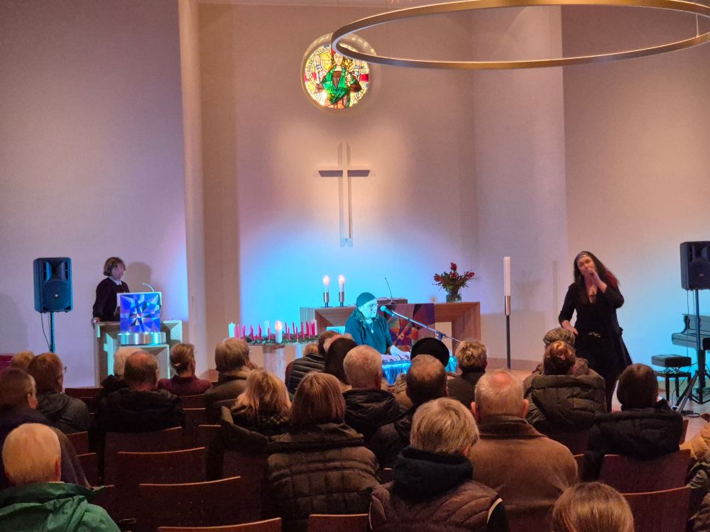 Die Band It's M.E. (Martina Maschke, Gesang, und Ecki Hüdepohl, Klavier) spielen beschwingt fetzig-besinnliche Musik in einer Kirche. Es sind viele Leute da. Im Hintergrund steht ein Wichernscher Adventskranz, bei dem die erste Kerze brennt, sowie unsere Pastorin.