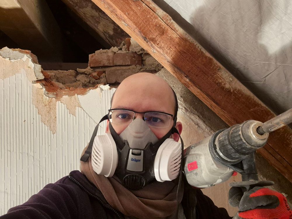 Selfie mit Maske und Bohrhammer vor Dachsparren und halber Mauer 