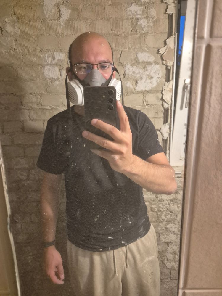 Spiegelselfie, ich mit Maske und besprenkelt mit Kalkputz vor einer rohen Ziegelwand 