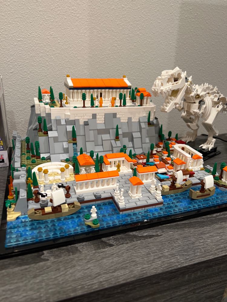 A picture of a Lego-like set version of the Acropolis in Athens. A Lego T-Rex is standing beside it, ready to eat the city.
