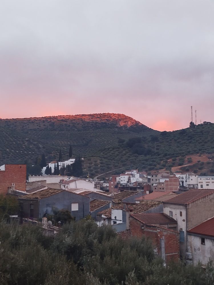 Panorámica de un atardecer donde se ven montañas con olivos en tonos rosados y casitas bajas de pueblo de color blanco