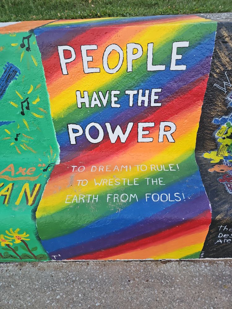A concrete wall bench. 2 foot wide sections get painted by ground snd individuals once a year. This is a photo of one section with a vibrant diagonal rainbow. Text is painted over it. 

"PEOPLE
HAVE THE
POWER

TO DREAM! TO RULE!
TO WRESTLE THE 
EARTH FROM FOOLS!"