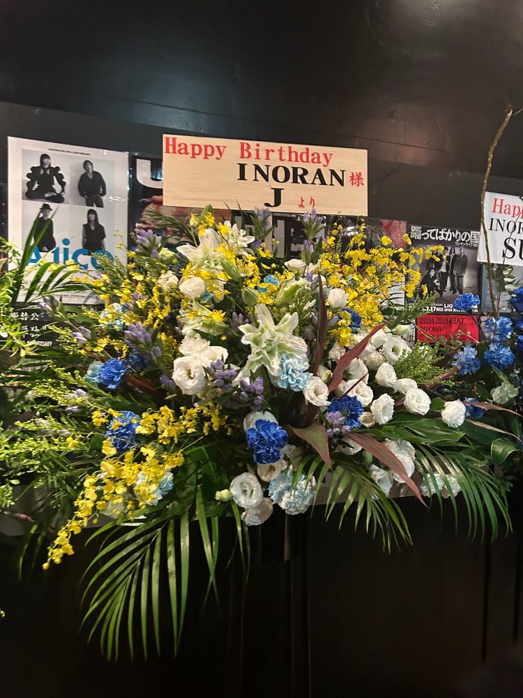 白い花メインに黄色・青い花がサブカラーのフラワースタンド
「Happy Birthday INORAN様　Jより」の白いカードがついている
