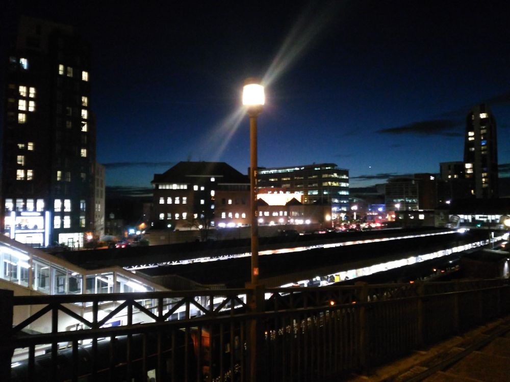 The lights of a town's railway station, office blocks and one of its lamp posts beaming into the dark blue of the night sky.