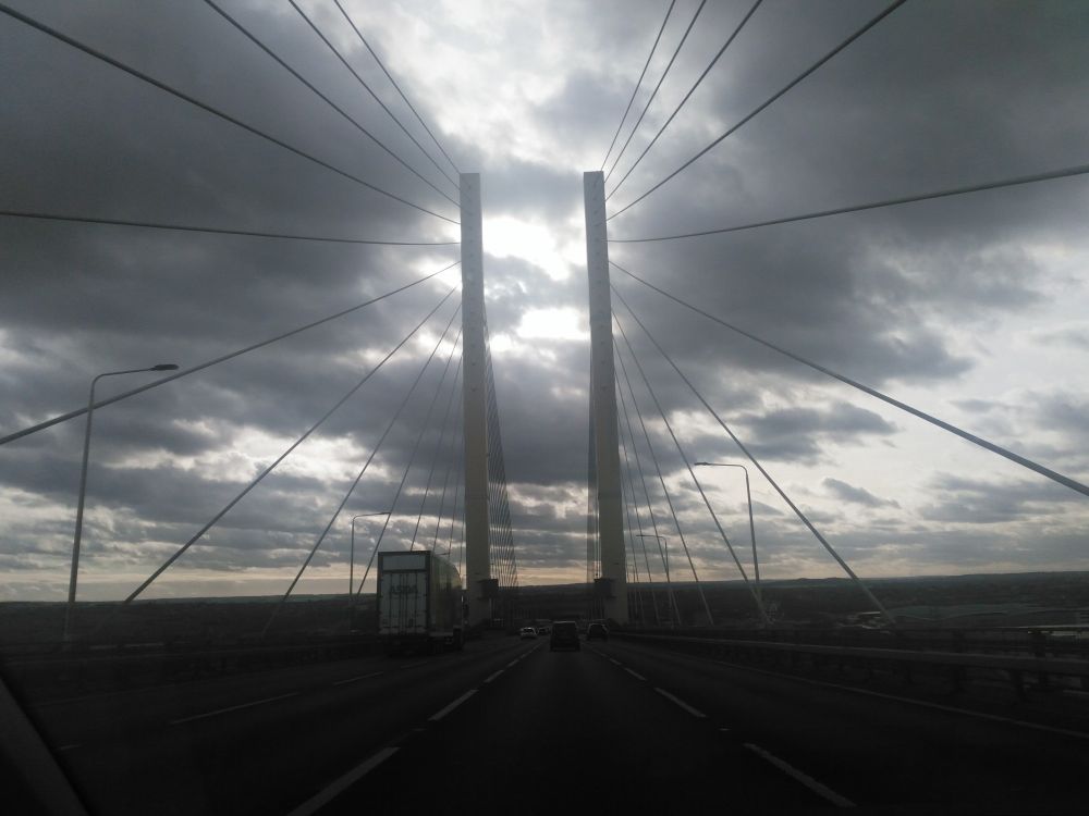 Sunbeams through grey clouds, shining between the suspenders of a bridge.