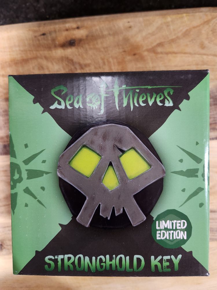 A green and black Sea of Thieves branded box, with the label "Stronghold Key" and displaying a picture of the contents, a small, black circle with a silver skull with green eyes