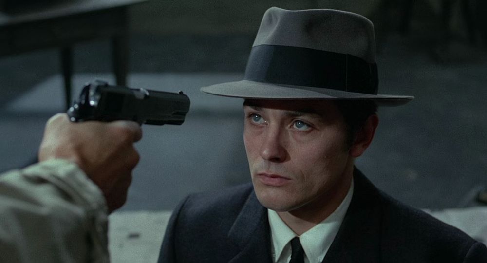 screencap from "le samourai" (1967 film) showing a man with clear blue eyes wearing a suit and hat looking at someone pointing a gun at his face; the holder of the gun is mostly off-camera, with just the hand and part of the arm visible, barrel of the gun barely in focus