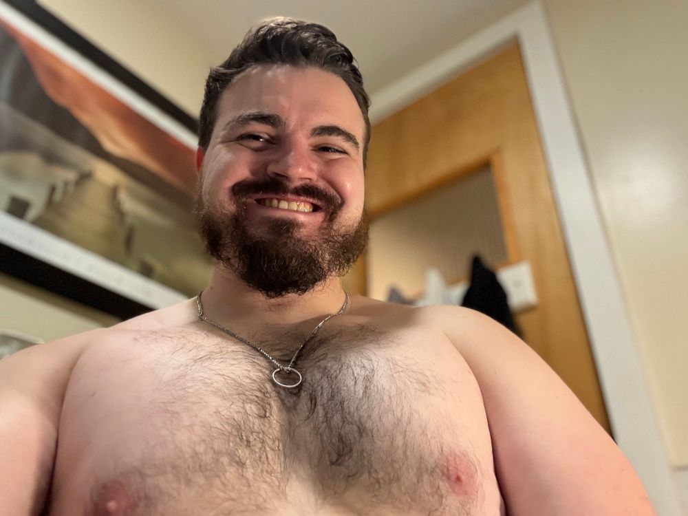 Man with brown hair and a beard wearing a cheesy grin, no shirt, and a silver necklace with a silver ring on it. By the most objective of judgements, his pecs are rather large