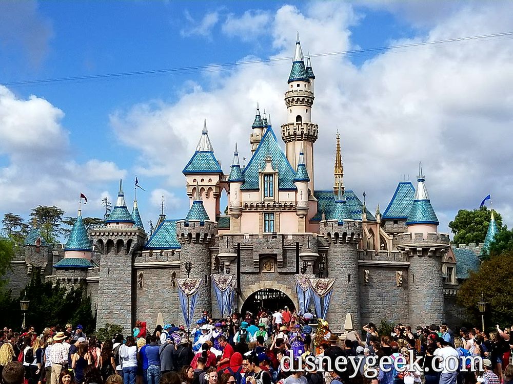 An image of Sleeping Beauty Castle at Disneyland, California from 2017. The shiny roof decorations from the 60th Anniversary Celebration in 2015 had inadvertently damaged the roofs of the castle, so they had to remain in place despite the rest of the 60th decorations being removed. The roofs are all outlined in a shiny blue metal and the tips of the castle spires resemble the diamond-shaped spires from the celebration. A crowd of people are seen below the castle, waiting to cross the drawbridge into Fantasyland.