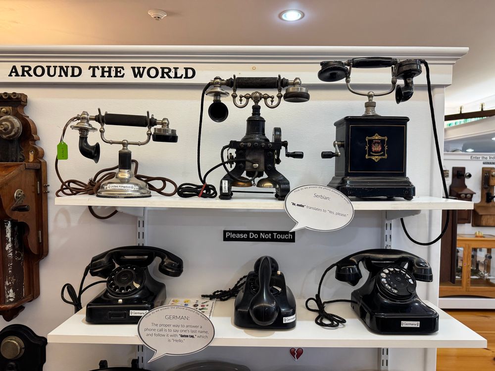 A display of six telephone headsets from the mid-20th century under the heading “Around the World”. All six feature a separate base and handset, and are made out of black Bakelite.