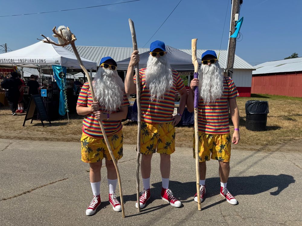Three persons dressed identically in blue caps, sunglasses, striped shirts, yellow printed Bermuda shorts, crew socks, and red converse. They all carry tall wooden staffs and wear long white costume beards and mustaches 