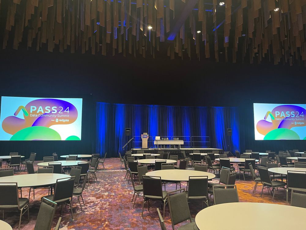 Large empty ballroom with tables and chairs set out in front of a stage with two large screens either side of the stage.