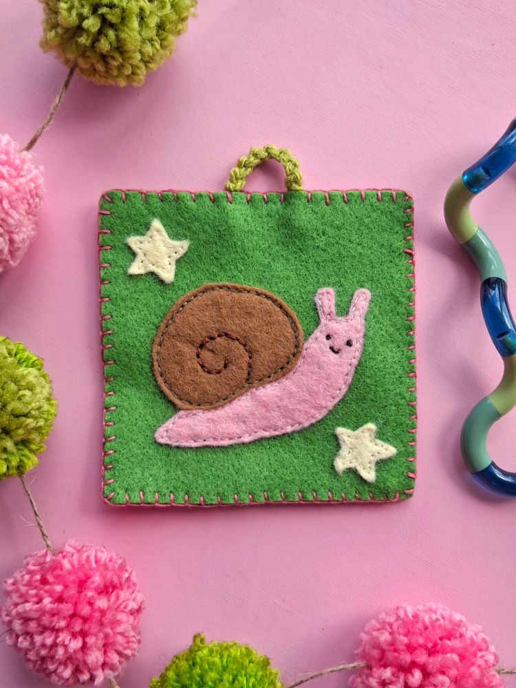 A photo of a small felt wall hanging, the piece shows a happy pink snail with a brown shell. There are two stars around the snail and they are on a green square of felt that has been blanket stitched around the edges. Behind the wall hanging the background is light pink with green and pink pom-poms on a piece of string. 