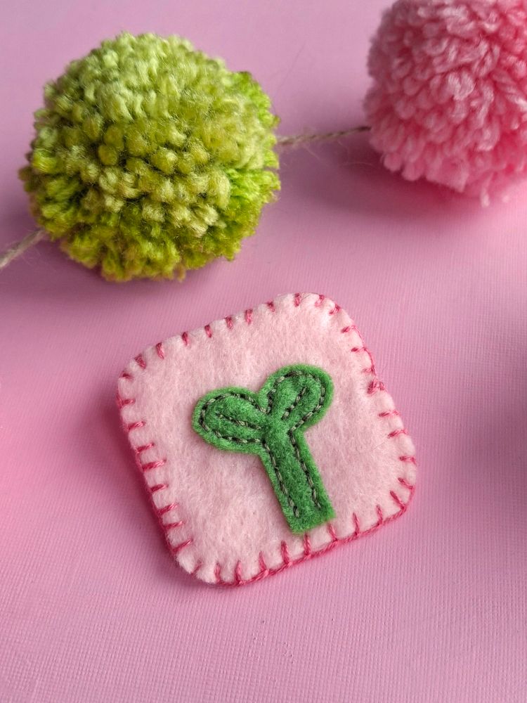 A photo of a pale pink felt pin badge of an embroidered plant sprout. The edge of the square badge is blanket stitched with darker pink thread, the background of the image is pink with green and pink pom-poms on a piece of string. 
