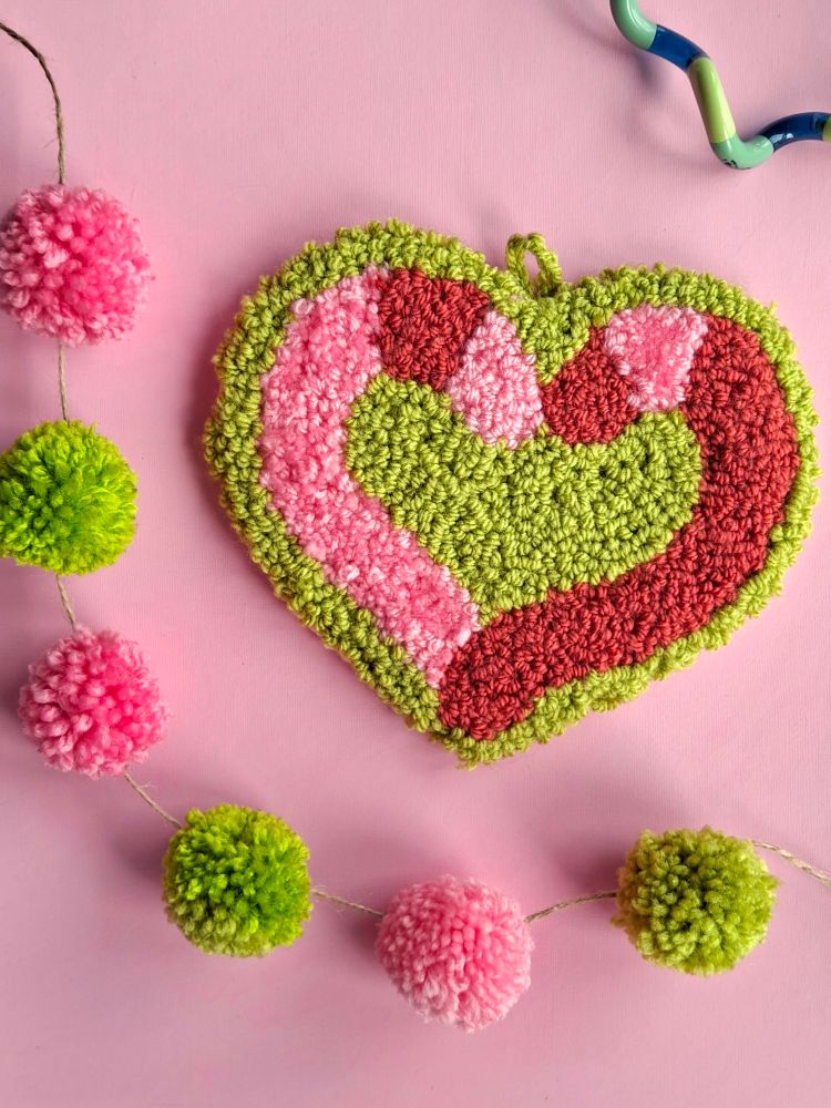 A photo of a punch-needle embroidery piece depicting two pink and red worms forming a heart shape on a green heart shaped background. Behind the piece, the background is light pink with green and pink pom-poms on a piece of string. 
