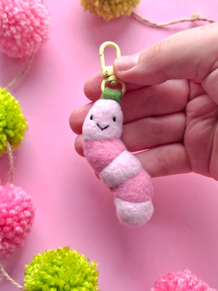 A photo of a hand sewn fleece worm keyring, the worm has a happy face and is attached to a yellow keyring clasp with a piece of green felt. The background of the photo is pink and the keyring is being help by a pale hand. 