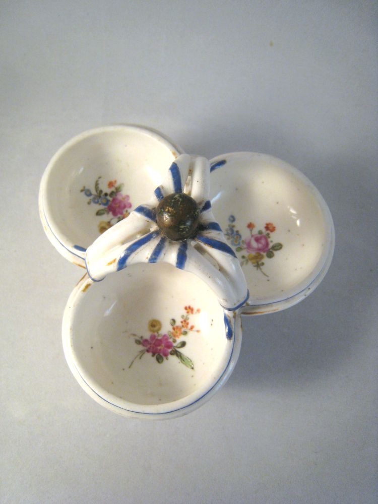The earliest known Sèvres example is from 1759, a form designed as a small basket with ribbons wrapped around a ball handle. This roughly-executed Mennecy or Bourg-la-Reine version is a later copy of that triple salt cellar form, but with only a trace of gilding due to the Sèvres royal monopoly on gilding wares at that time.
