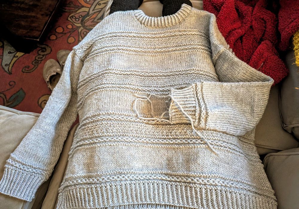 A grey hand knit sweater with loose yarn pieces hanging from the sleeve. It is draped across a woman's lap.