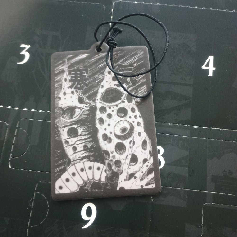 A rectangular air freshener with Junji Ito's art (some kind of thing covered in holes with eyes peeking through and what appears to be part of a caterpillar)