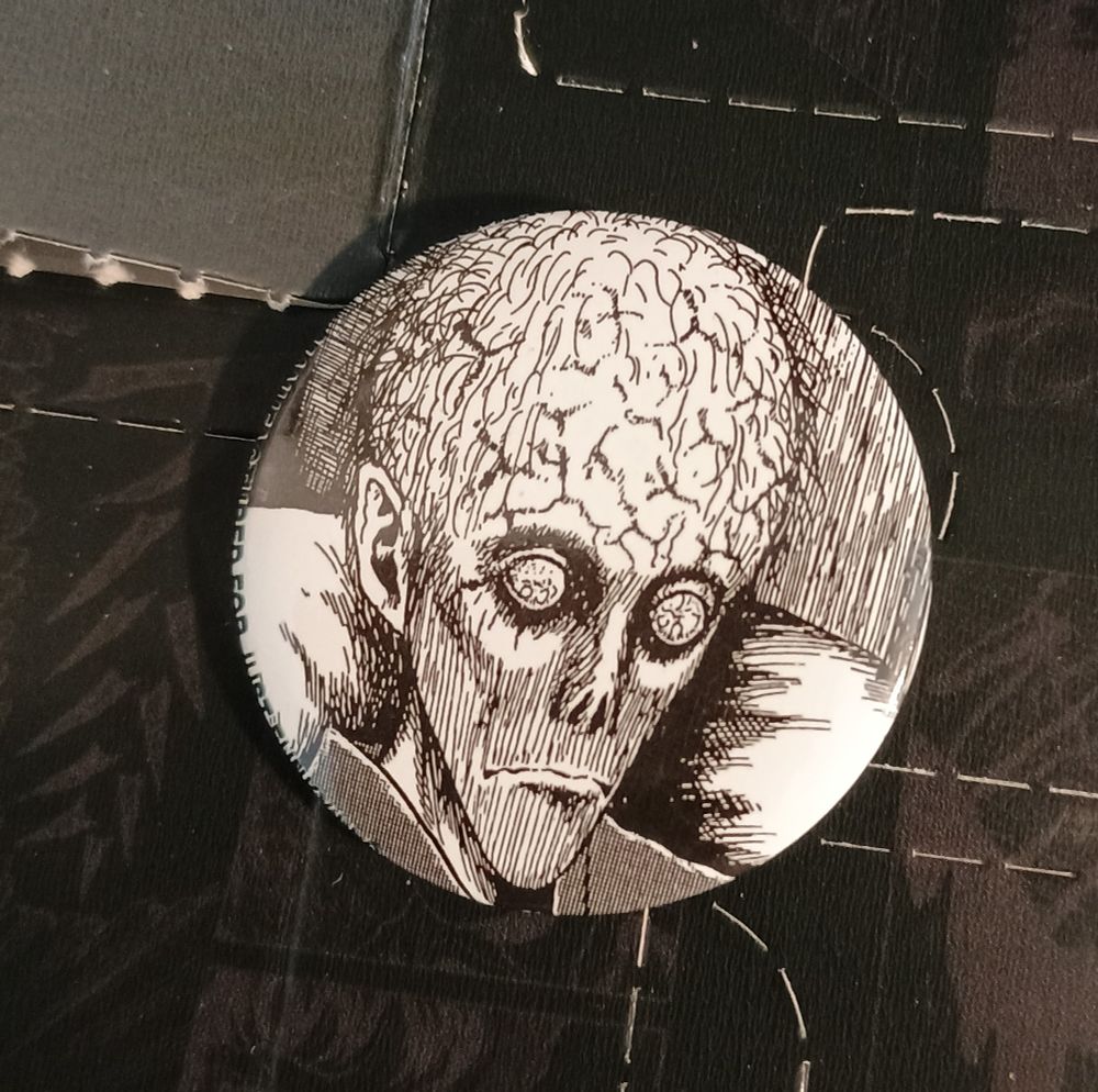 A round button depicting Junji Ito's art of a gaunt man with his head all big like an alien's, covered in brain wrinkles and sparse hairs. His bloodshot eyes are bulbous and without irises and pupils.