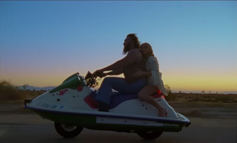 Sabrina Carpenter with a man on a jetski riding it like a motorcycle down a highway at sunset